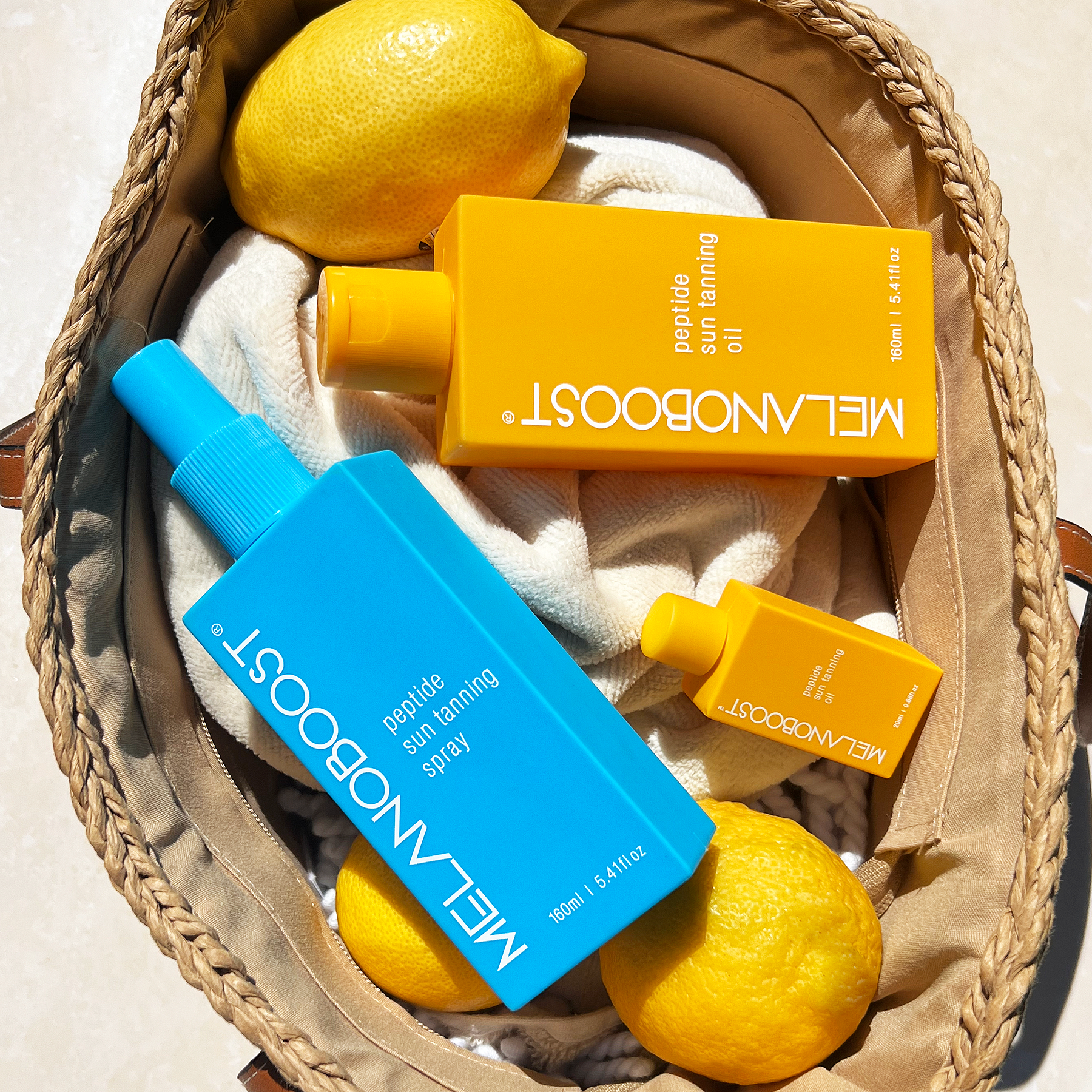 Melanoboost Peptide Sun Tanning Oil and Spray bottles in a woven basket with lemons.